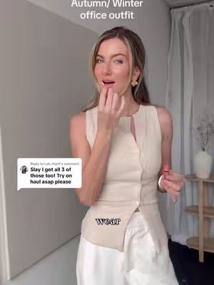 A woman stands in a well-lit indoor space. She is wearing a cream-colored top and white pants. The background is a light color and the overall composition is a medium shot.