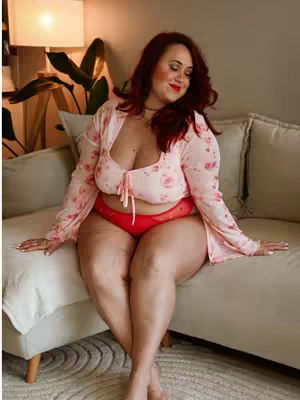 A woman with long red hair sits on a beige couch wearing a floral patterned, light pink top and matching red underwear. She is smiling while looking down. The background includes a lamp with a warm light, a green plant, and neutral-toned pillows.