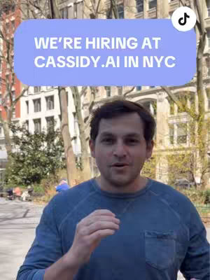 A man in a dark blue sweater is standing in an outdoor setting. A light blue rectangular text overlay is positioned above him, featuring white text. The background consists of a building and trees.