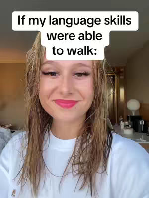 A selfie-style shot of a woman with wet blonde hair and a white t-shirt. Text appears above her head in a white rounded rectangle. She is smiling with dark eye makeup and vibrant pink lipstick. The background is blurred, appearing to be an interior space with warm lighting.
