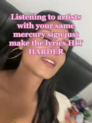 A selfie-style video showing a young woman looking slightly upwards, smiling, and with text overlayed on the image. The background is blurred, but includes a dark-colored surface and some items that are out of focus.