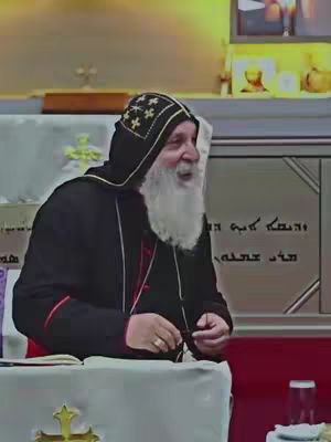 An older man with a white beard and a black hood is seated, speaking to an unseen audience. He wears a black robe with red accents and has a gold ring on his finger. The setting includes a cross on a table and text in a foreign language on the wall.