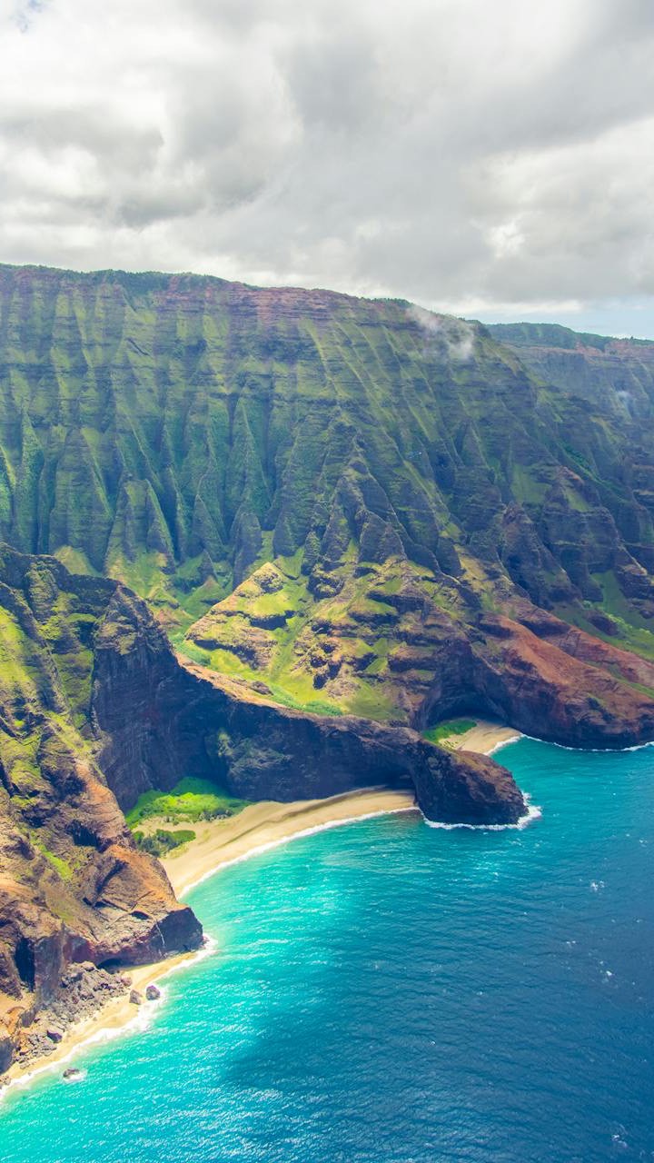 An aerial view of a lush, green, mountainous coastline meeting a vibrant turquoise ocean and sandy beach. The sky is cloudy. The composition is divided diagonally. The top left is sky, the middle is a rugged mountain cliff face, and the bottom right is the ocean and beach. The colors are bright and saturated, with greens, blues, browns, and whites creating a scenic view.