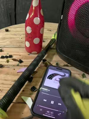 The cover image shows a wooden table with a red and white polka-dotted bottle, a broken glass, a black speaker with a red center, and a smartphone displaying music. A black-handled object, possibly a golf club or similar, rests horizontally across the frame. The smartphone is on the right side, next to the speaker, and the phone's screen shows music with the play button in the center. The color scheme is primarily wood tones contrasted with bold red, black, and the phone's interface.