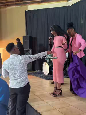 A man in a printed shirt points his hand towards a woman in a pink dress, with another woman in a pink jacket standing nearby. The background shows a stage setup with a drum set and black curtains. The text overlay reads "The anointing is transferable."