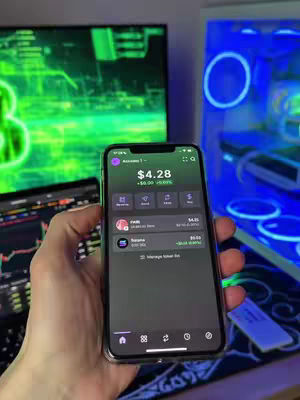 A hand holding a smartphone with a financial app displayed on its screen, set against a backdrop of computer equipment and a neon-lit blue computer case. The phone screen shows account details with a financial summary and app icons. Behind the hand, a monitor displays what appears to be a stock chart.