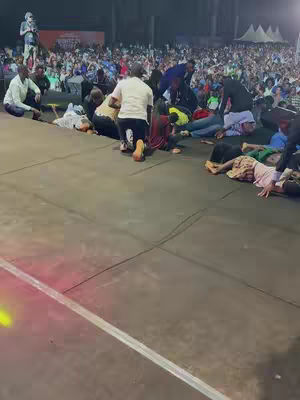 A wide shot depicting a large outdoor gathering, likely at night. The foreground is dominated by a stage or elevated platform where several people are kneeling or lying down. The background is filled with a large crowd of people, possibly spectators, with tents visible on the far right. The scene is illuminated, likely by stage lights and ambient light from the event. The overall colors seem dark, with the lights creating contrasting spots of brightness.