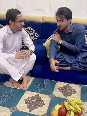 Two men are seated on a blue sofa, engaged in conversation. One man is dressed in a white outfit and glasses, while the other wears a blue traditional garment. A fruit bowl with bananas and other fruits is placed in front of them.