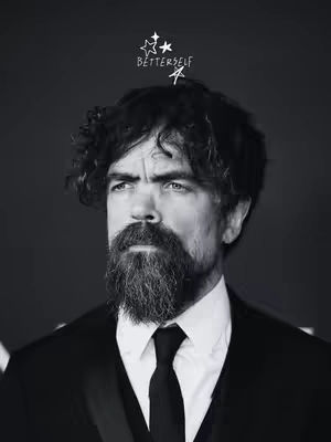 A black and white portrait of actor Peter Dinklage, facing left with a serious expression. He is wearing a black suit, white dress shirt, and a black tie. He has a long, full beard and curly hair. Above his head, there is a small graphic with stars and text that says 'BETTERSELF'.