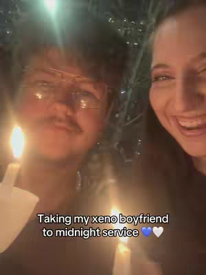 A selfie featuring two people in a dimly lit setting, likely at a religious event. A man with glasses and a mustache on the left holds a lit candle, while a smiling woman on the right is also holding a candle. Text below says "Taking my xeno boyfriend to midnight service."