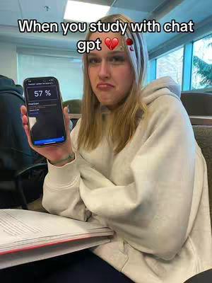 A young woman is holding a black smartphone displaying a user interface with a percentage of 57%. She has a confused expression, with a slightly furrowed brow and a downturned mouth. She is wearing a light-colored hoodie and has blonde hair, and she is sitting in a classroom setting. The background includes desks, chairs, and windows letting in natural light. The text overlay says 'When you study with chat gpt' and has a heart icon and flowers next to it