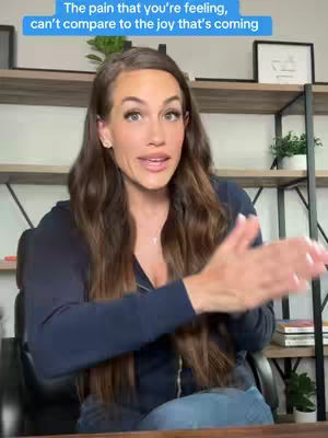 A woman with long brown hair is in front of a bookshelf and wooden background. She is wearing a dark blue top, and speaking with her hands up. There is text overlay above her head in a light blue box