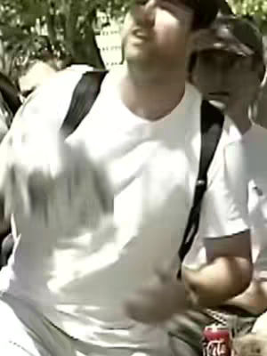 A man with a white t-shirt and black backpack straps is at the front of the image, appearing to be talking animatedly. Other people are visible, and a Coca-Cola can is visible. The background is out of focus.
