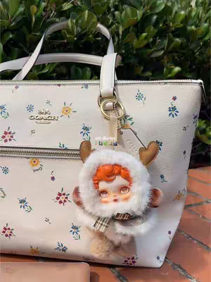 A purse with small floral patterns is the primary focus, with a charm of a doll-like figure hanging from the handle. The doll has orange hair, and wears a furry cap and scarf, and has an overall cute appearance. The background appears to be a garden scene.