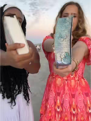 Two young women are positioned in front of a beach scene, holding up books to the viewer. The woman on the left is wearing a white head band and holding a book or journal with blank pages, while the woman on the right holds an open blue book with butterfly illustrations. The backdrop shows a beach during what appears to be a sunset with hues of pink and orange. The two women appear to be out doing a photoshoot.