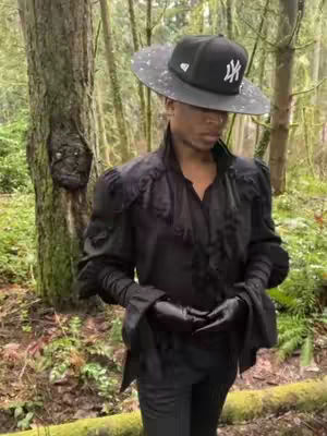 A person wearing a hat and all-black clothing poses in a forest. The person is facing slightly down, with their hands clasped in front, the hat obscures the eyes. Behind the person, there are trees in the background .