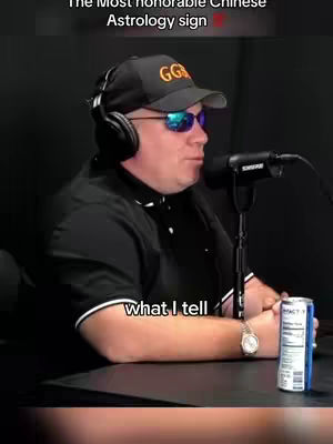A man wearing a black cap, sunglasses, and headphones speaks into a microphone while seated, with a can visible beside him.