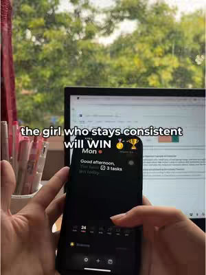 The image shows a person holding a phone with an app open. The phone is displaying a task list with three tasks marked as incomplete. Behind the phone is a laptop with a document open. The background is a blurred view of outdoor foliage, suggesting a window setting. Text is overlaid at the top, highlighting the video's motivational theme. The overall composition includes natural lighting, showcasing a motivational theme centered on productivity and consistency.