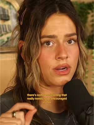 A close-up shot of a woman looking directly at the camera with a concerned expression. She has shoulder-length wavy brown hair, styled with a couple of small pink hair clips. Her eyes are brown, and she wears a dark gray top. Her hand is raised and she is holding a microphone.