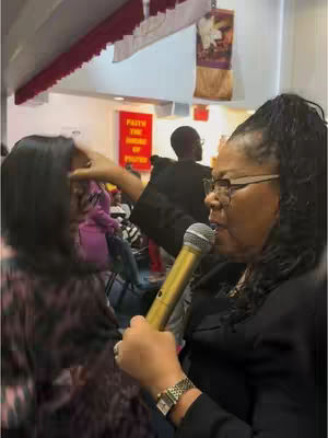 An image of a woman in glasses holding a microphone in a church, touching a woman on the head.
