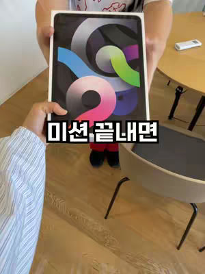 A person holding an iPad in its box, presenting it towards the camera. Another hand can be seen in the bottom left of the frame. The background consists of a wooden table and a beige chair. The text overlay '미션. 끝내면' (If you finish the mission) is placed over the iPad and its holder.
