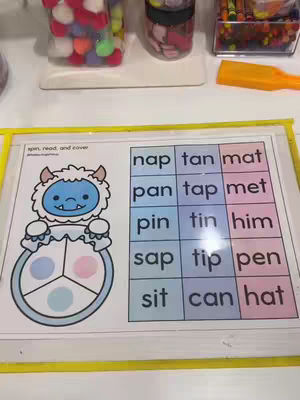 An image showing a kindergarten activity. It features a printed sheet with a cartoon monster holding a spinner wheel and blocks with short words for the kids to read and cover. The background shows school supplies