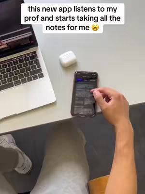 A top-down shot shows a person's hands interacting with a smartphone sitting on a gray desk. Beside the phone is a laptop with its screen open, and a pair of white earbuds. The person's legs and shoes are visible in the lower part of the image. The focus is on the phone and laptop, suggesting a tech-related theme.
