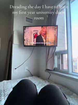 The image shows a person's legs resting on a patterned bedspread, with a large window and a TV mounted on a wall visible in the background. The TV is displaying a scene, and the text overlay says, 'Dreading the day I have to leave my first year university dorm room.'