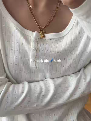 The image shows a close-up view of a person's upper chest and neck. They are wearing a white, long-sleeved shirt with a textured, textured pattern. A gold necklace is visible around the neck, with a small, decorative pendant. The text overlay 'Primark pjs' is present, along with a sleeping face emoji and a cloud emoji
