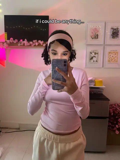 A woman is shown in a mirror selfie, holding a smartphone in front of her face, obscuring her features. She wears a light pink long-sleeved shirt and cream-colored sweatpants. She has curly hair styled with a pink headband. The background includes a wall with decorative framed art, a shelf with decor, and part of a mini-fridge. The lighting in the room is a mix of pink and natural tones, creating a soft ambiance.