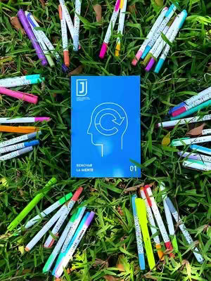 A top-down shot of a patch of green grass with numerous colorful pens scattered around a blue booklet. The booklet is in the center, featuring a white line drawing of a head with a recycling symbol inside. Text is visible on both the booklet and the pens. The image is well-lit and has a natural, slightly out-of-focus background.