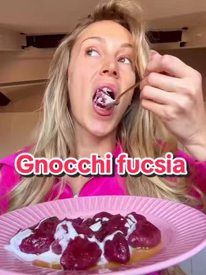 A woman with long blonde hair is shown from the chest up, looking upwards with her mouth open as if tasting food. She is wearing a pink zip-up top. In front of her is a plate of pink gnocchi with white sauce.