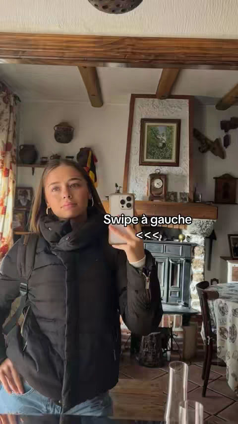A young woman holds a smartphone, taking a selfie in a domestic setting. She stands in front of a large mirror. Behind her, a stone fireplace, framed artwork, and other decorative elements suggest a cozy, lived-in space.