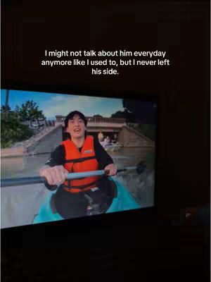 The image shows the blurred screen of a television displaying a man in an orange life vest in a boat. The man seems to be rowing in a body of water. The television is set against a dark background, with text overlayed at the top.