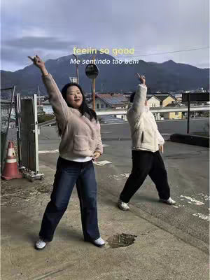 Two people, one wearing a light-colored fuzzy jacket and jeans, and the other wearing a white jacket and black pants, stand on a street with their arms raised towards the sky. Text overlays are present on the image. The background includes a mountain range and buildings.
