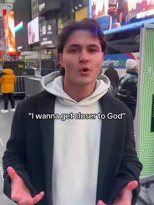A man in a white hoodie and black coat stands in front of brightly lit billboards with text overlayed. He's looking at the camera with his hands open, seemingly inviting viewers. The background has figures of people, with a sign.