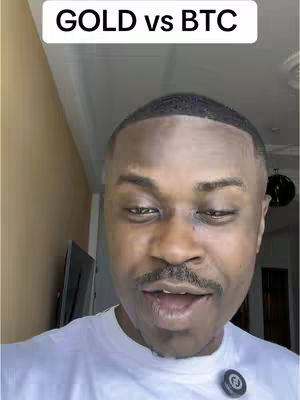 A man with a short haircut and a mustache is looking at the camera with an open mouth, in front of a gold-colored wall. White uppercase text "GOLD vs BTC" appears in a white rectangle at the top of the image. The man wears a white t-shirt. Part of a television can be seen behind the man. He has a black circle badge object on his shirt.