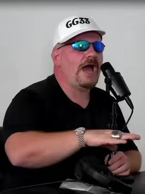 A man is facing the camera, holding a microphone, and wearing a white baseball cap with the letters GG33 on it, sunglasses, and a black shirt. His mouth is open, and one hand is raised with a ring on the index finger. The background is plain.