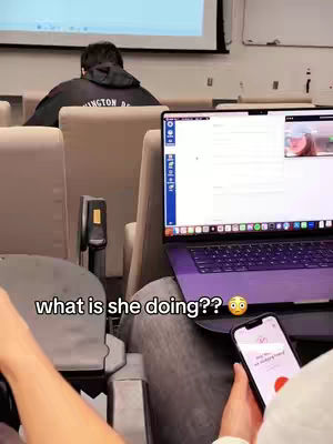A person is sitting in a classroom setting with a laptop open, and another person is sitting behind them. The focal point is a phone held in the foreground, displaying a screen with a red microphone icon. The text overlay is visible on the image: what is she doing??