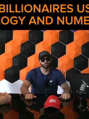 A man wearing sunglasses and a baseball cap sits at a table with a red hat and two microphones. Behind him is a patterned wall of orange, black and chrome-colored panels.