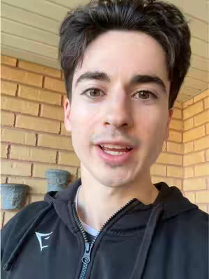 A selfie-style shot of a young man indoors. He is positioned in the foreground, looking towards the viewer and speaking. The background consists of a tan brick wall and the edge of a white surface on the right. A couple of gray flower pots are visible, seemingly hanging from the wall.