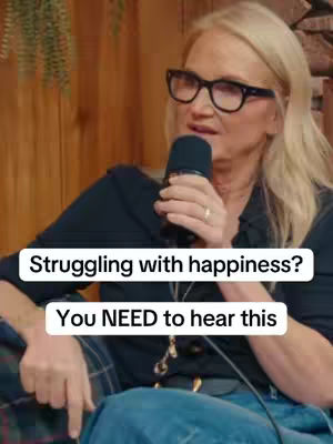 A woman wearing glasses and holding a microphone is speaking. Above her, in a white box, are the words "Struggling with happiness?". Below, also in a white box, are the words "You NEED to hear this". The background is a blurred wooden seated area.