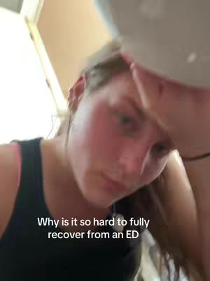 A slightly upward-angled selfie shows a young woman with fair skin and reddish cheeks, possibly due to blushing or a medical condition. Her expression is downward and thoughtful, her eyes casting a heavy gaze. The lighting seems to be from above, creating shadows on her face. The background is indistinct, with a glimpse of a white surface and another out-of-focus object. Her dark tank top is partially visible. Text overlayed in a clean white font reads "Why is it so hard to fully recover from an ED".