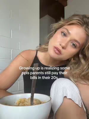 A young woman with blonde hair and fair skin leans towards the camera. She is wearing a black spaghetti-strap top. A bowl of food sits in front of her. Overlay text on the image reads "Growing up is realizing some ppls parents still pay their bills in their 20s".