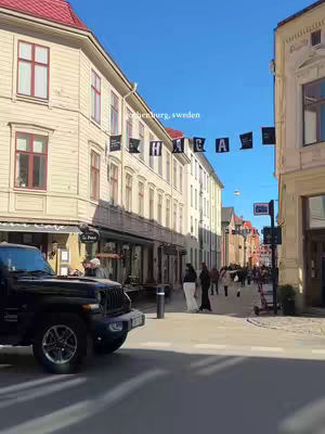 A street scene in Gothenburg, Sweden. The image shows a street lined with buildings on either side, with a black Jeep on the left. People are walking in the distance. Above the street, black banners with white text are strung across. The sky is clear blue.