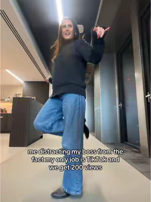 A woman poses in a hallway with one leg bent and arms raised, appearing to dance or gesture. She wears a black sweater, jeans, and flats. Text overlays at the bottom of the image provide context.