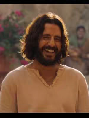 The image shows a man with brown wavy hair, a beard, and a gentle smile. He is wearing a simple, light-colored shirt, and the background is softly blurred, suggesting an outdoor setting with a hint of greenery and other figures. The lighting seems natural, highlighting his facial features.