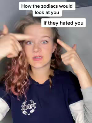 A young woman with light skin and reddish-brown curly hair is centered in the frame. She is wearing a dark blue sweatshirt with a white collar and sleeves, and a logo on the chest. The text overlay at the top reads "How the zodiacs would look at you" and "If they hated you." The woman is looking directly at the camera with a slightly concerned expression, and is pointing at the side of her head with both index fingers. The background seems indoors, with a plain light-colored wall.