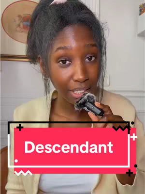 A woman with dark hair in a high ponytail is holding a microphone near her mouth. She is wearing a cream-colored blazer over a white shirt. A red rectangle with white text reading "Descendant" is overlaid on the image.
