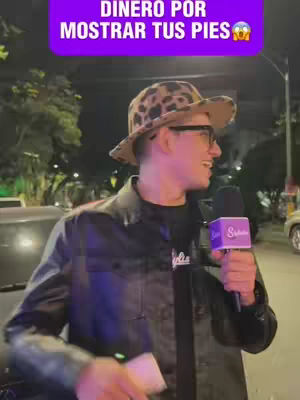 A man with glasses and a leopard-print hat is outside at night, holding a microphone, with a purple banner above him that says "Dinero por mostrar tus pies"